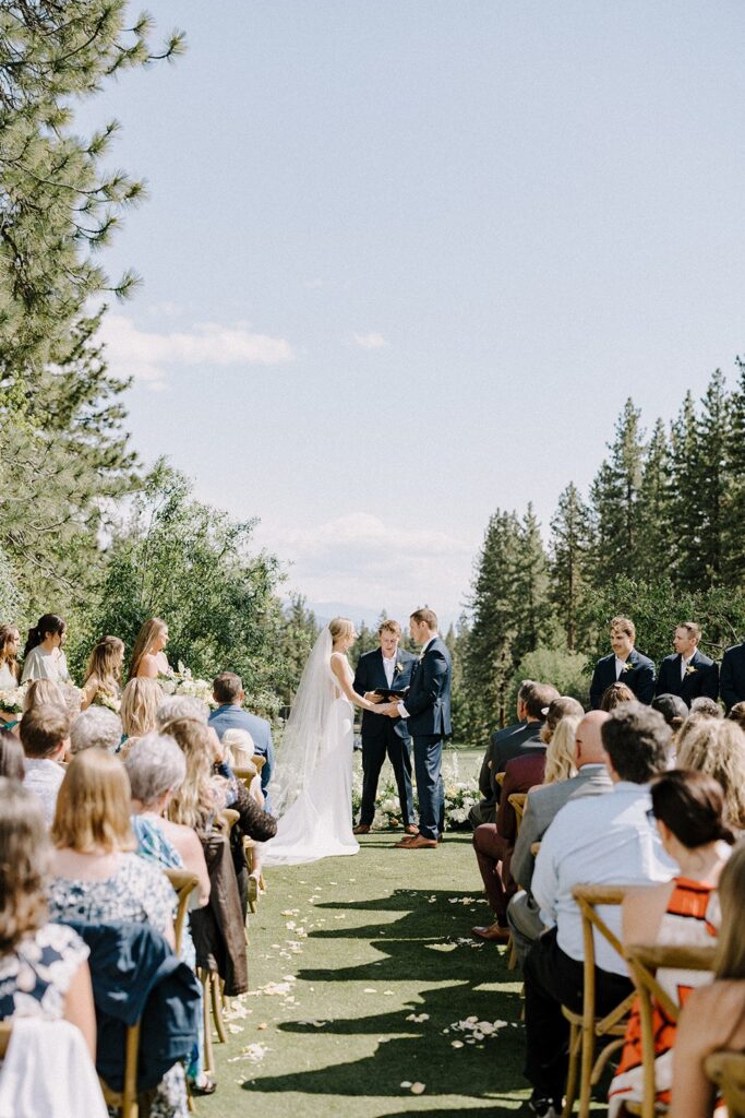 A couple stands before an officiant during an outdoor wedding ceremony, surrounded by guests seated in rows, with a backdrop of trees and a clear sky.
