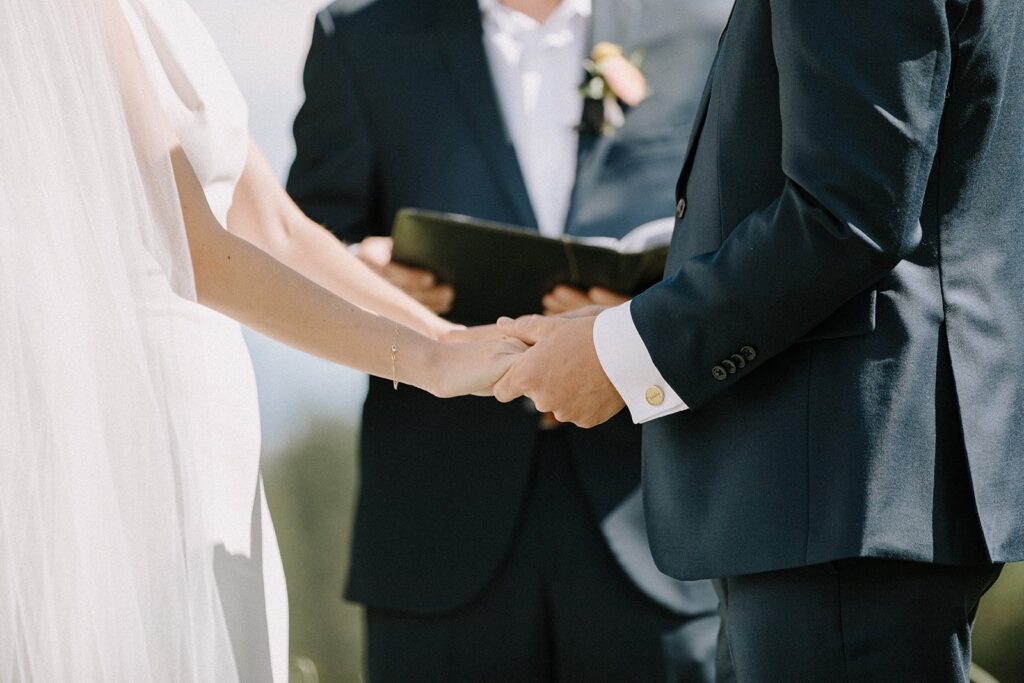 A bride and groom holding hands during a wedding ceremony, with an officiant in the background holding a book.