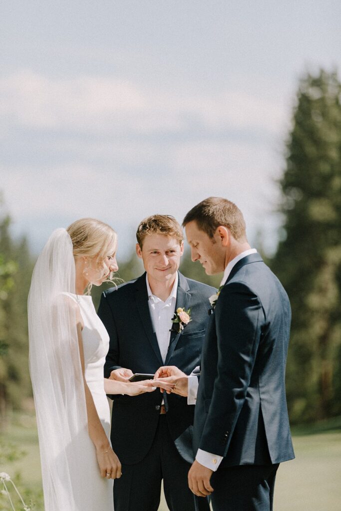 A couple is exchanging vows during a wedding ceremony outdoors with a person officiating. The bride is in a white dress with a veil, and the groom is in a dark suit.