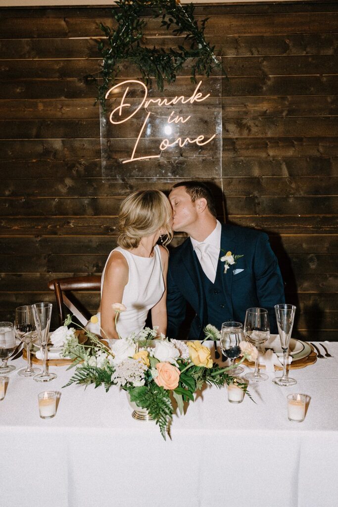 A couple kisses at a wedding reception table with a "Drunk in Love" sign behind them.