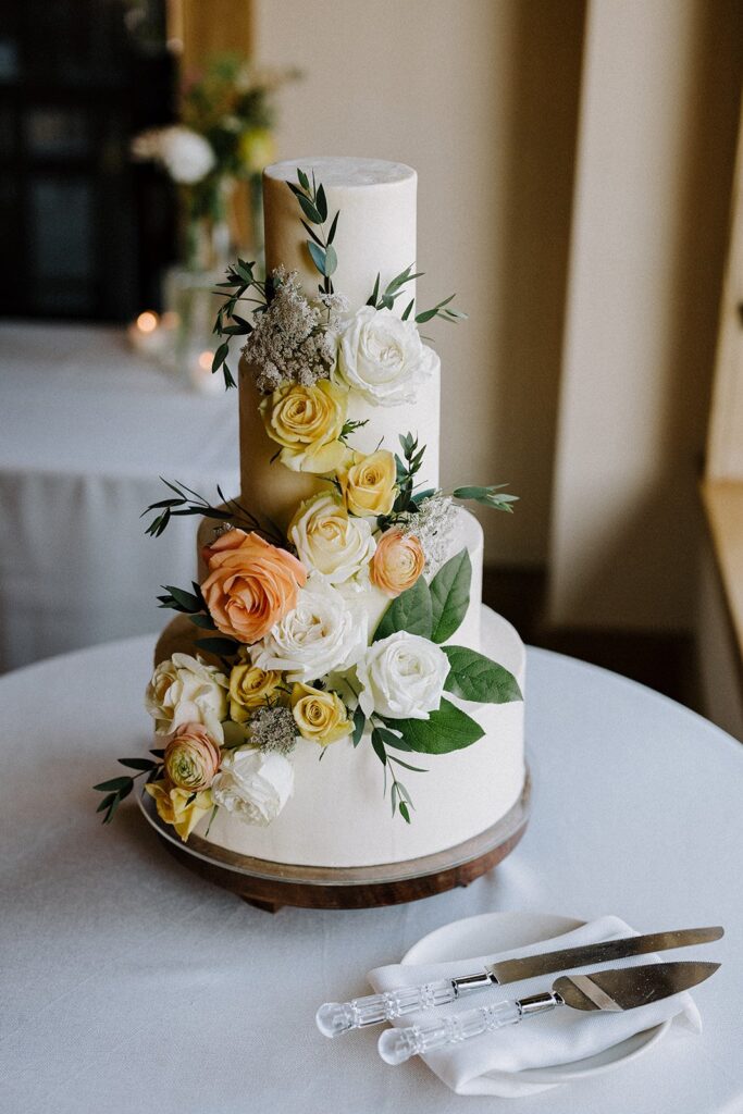 Three-tiered white wedding cake adorned with white, yellow, and peach roses, placed on a round table with two cake knives on a napkin beside it.
