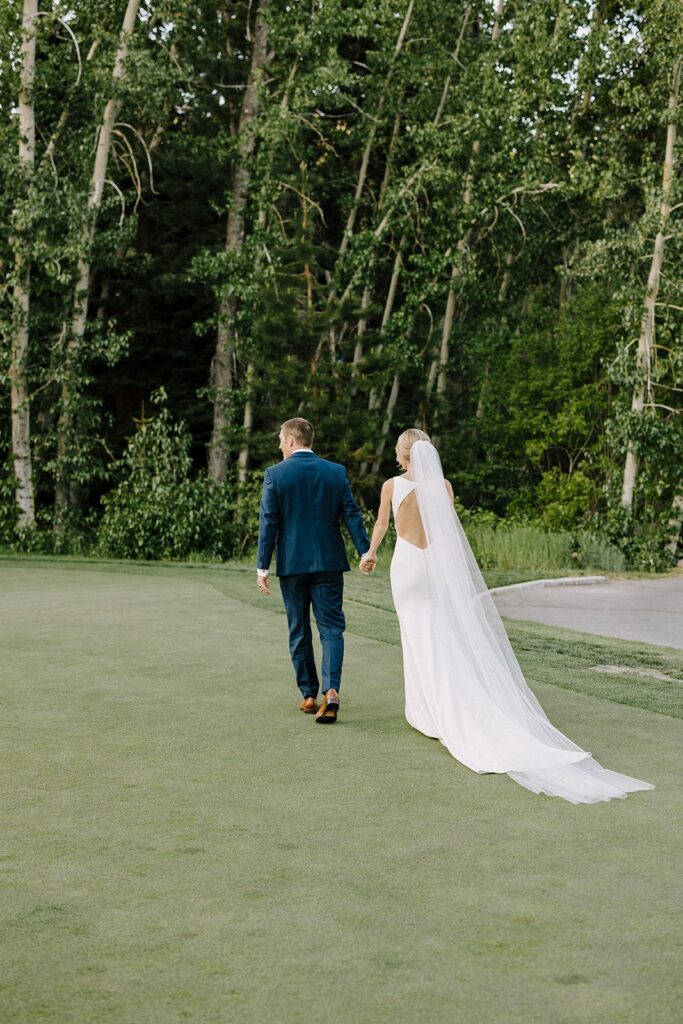 Bride and groom walk hand in hand on a grassy field, surrounded by tall trees. The bride wears a long white gown with a veil, and the groom is in a blue suit.