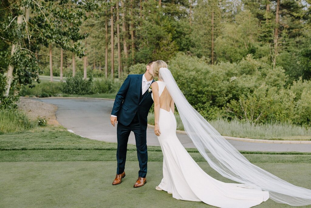 A couple in formal attire shares a kiss outdoors, with lush greenery and a path in the background. The woman wears a long, flowing veil.