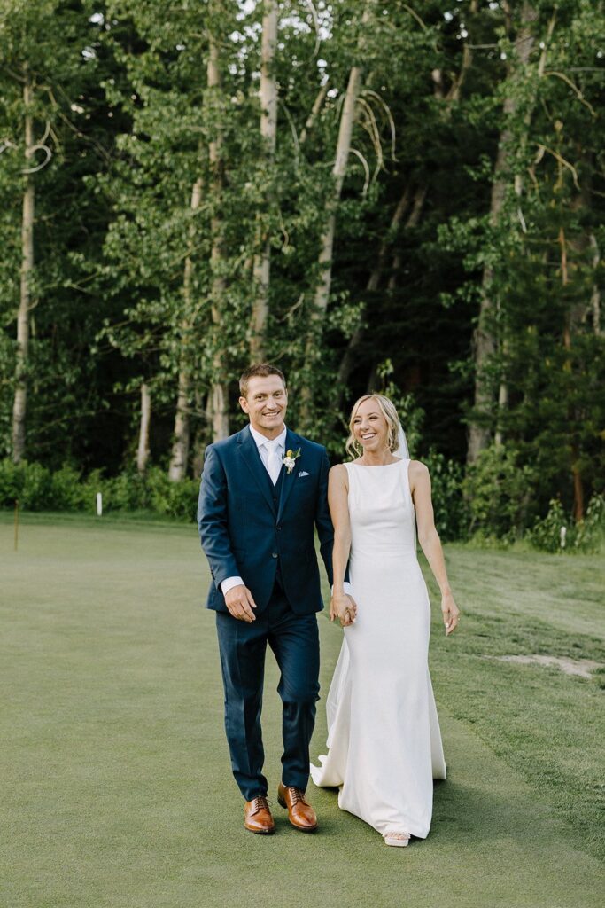 A couple in formal attire walks hand in hand on a grassy area, surrounded by tall trees.