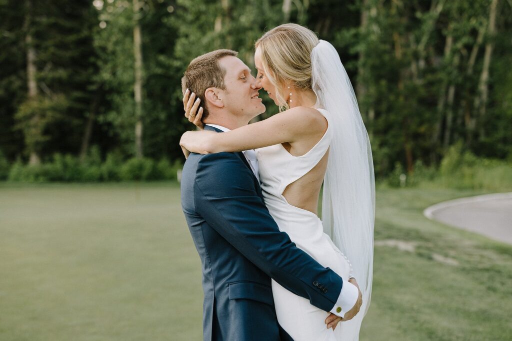 A bride and groom embrace and smile at each other outdoors, with green trees in the background. She wears a long white dress and veil, and he is in a blue suit.