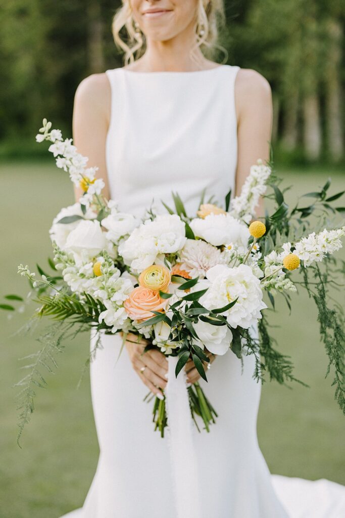 A woman in a white sleeveless dress holding a bouquet of white, yellow, and peach flowers with greenery.