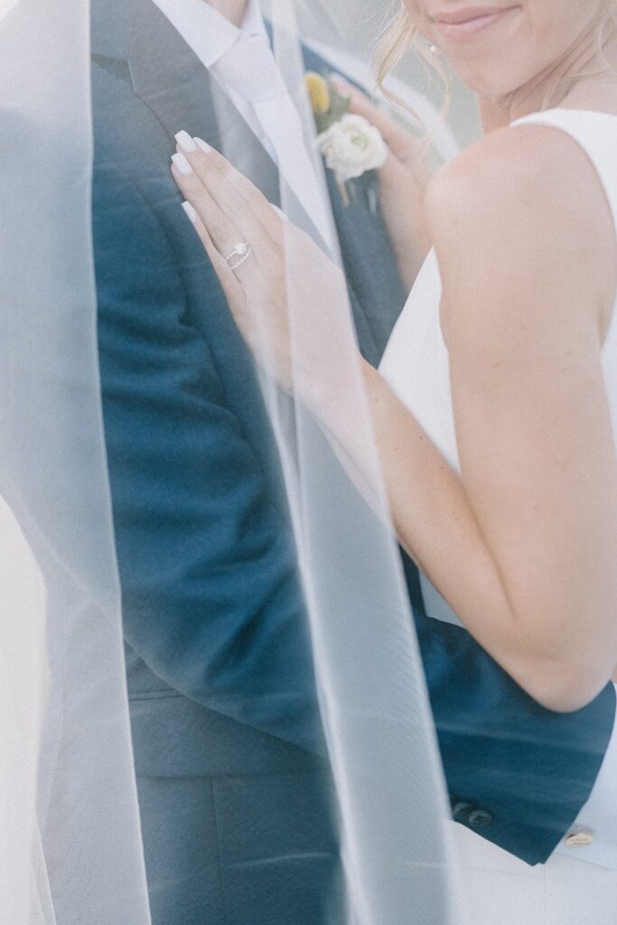 Bride and groom embracing, partially covered by a sheer veil. Bride's engagement ring and groom's suit are visible.