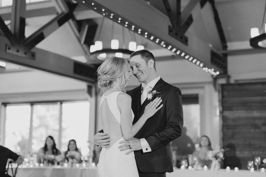 A couple in formal attire is dancing in a large hall with guests seated in the background.