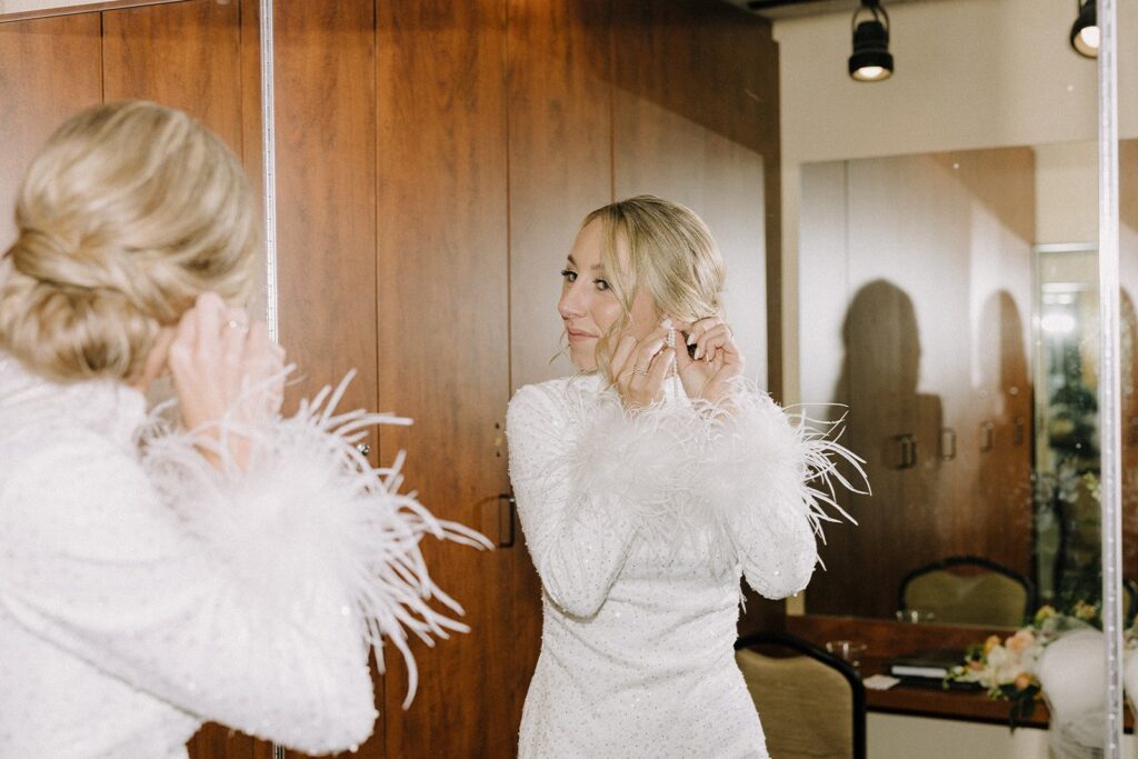 A person in a white dress with feather cuffs adjusts earrings while looking in a mirror.