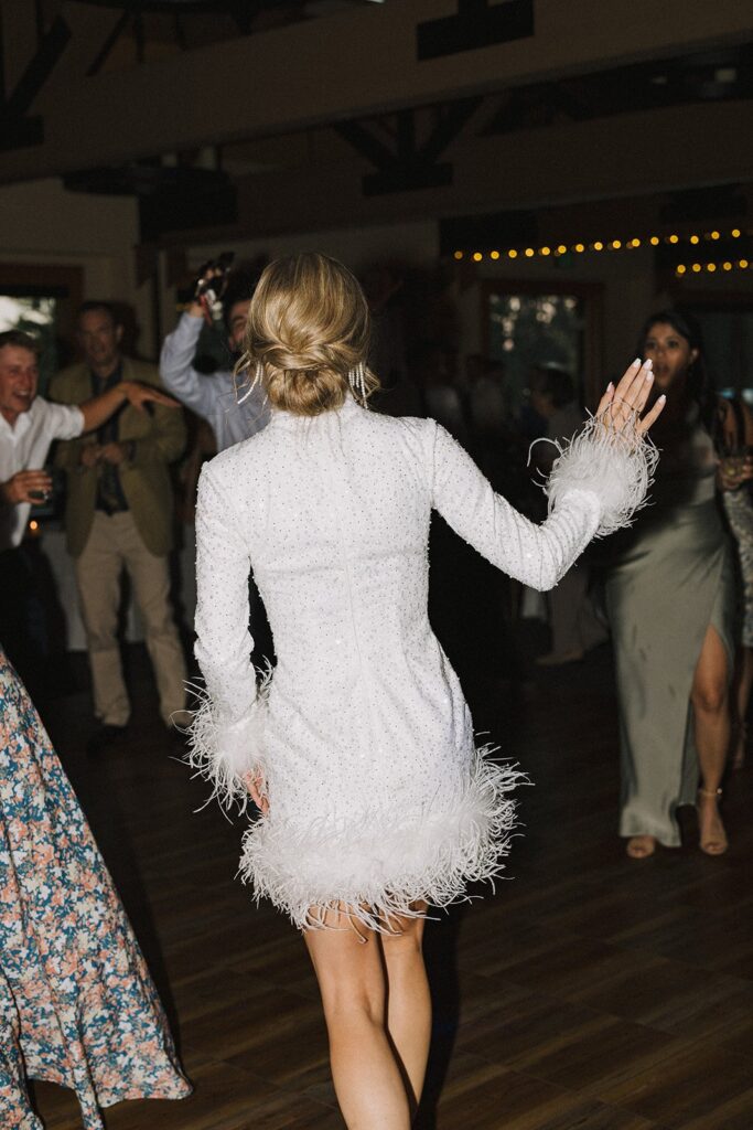 A woman in a white, feathered dress is dancing with her back to the camera. Others are dancing around her in a dimly lit room.