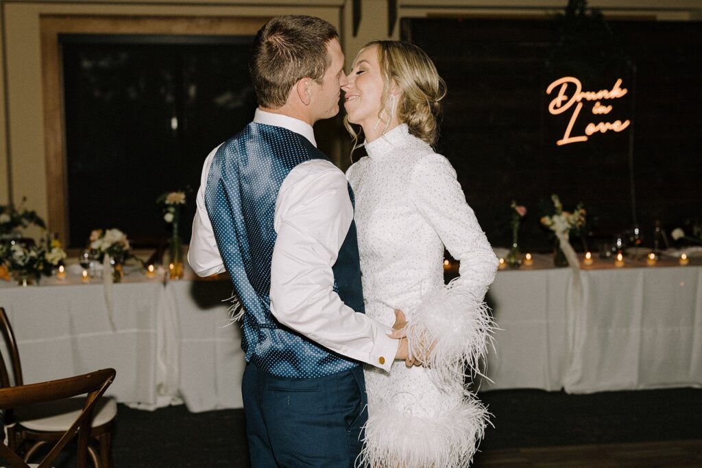 A couple dressed in formal attire dances closely in a dimly lit room, with a "Drunk in Love" neon sign and a decorated table in the background.