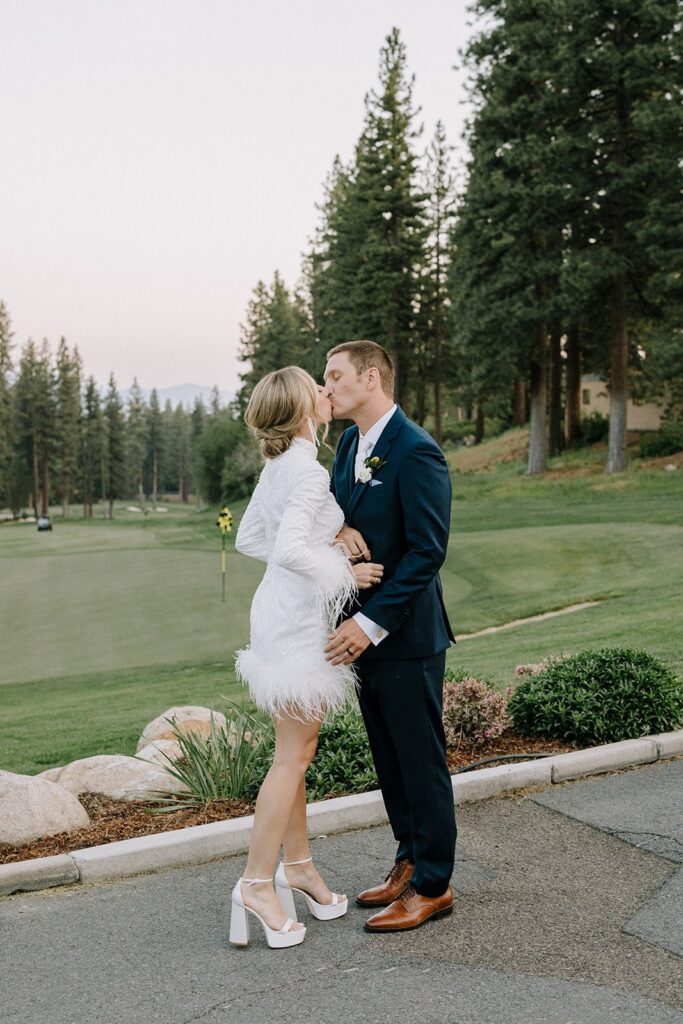 A couple kissing outdoors near a golf course, surrounded by trees. The woman is wearing a white dress with feather details, and the man is in a dark suit.
