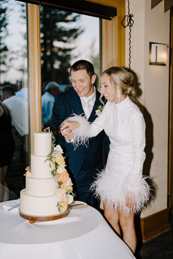 A couple in formal attire is smiling as they cut a three-tiered cake adorned with flowers at an indoor event.
