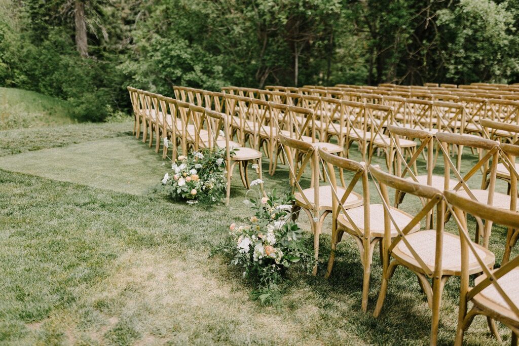 Outdoor wedding ceremony setup with rows of wooden chairs on a grassy area, surrounded by trees. Floral arrangements line the aisle.