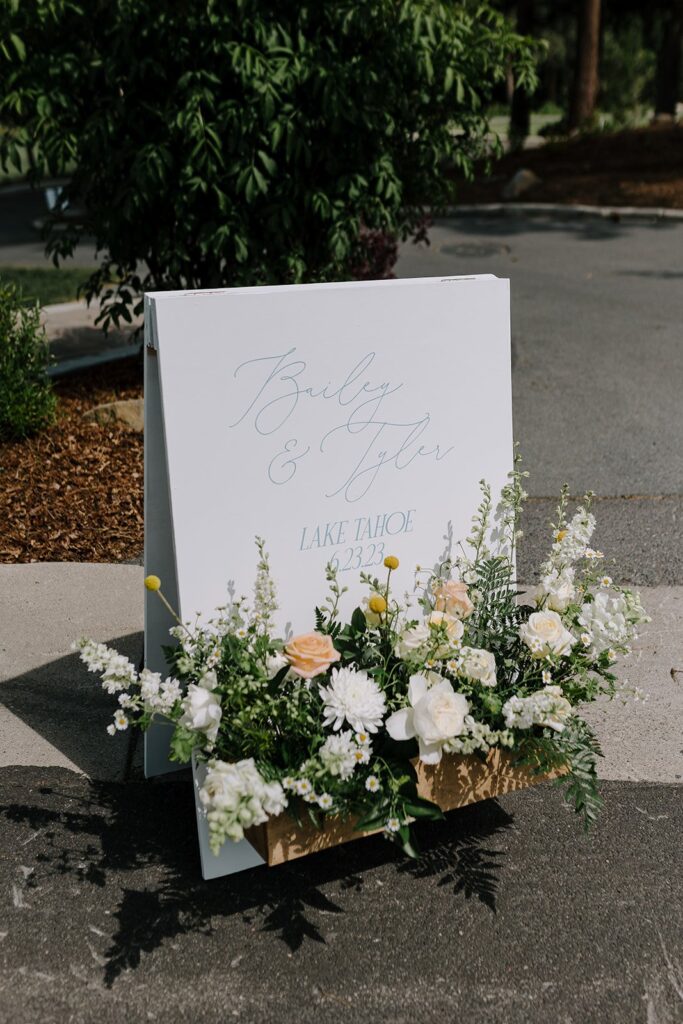A sign with "Bailey & Tyler, Lake Tahoe, 2023" stands on a path, adorned with white and peach flowers and green leaves.