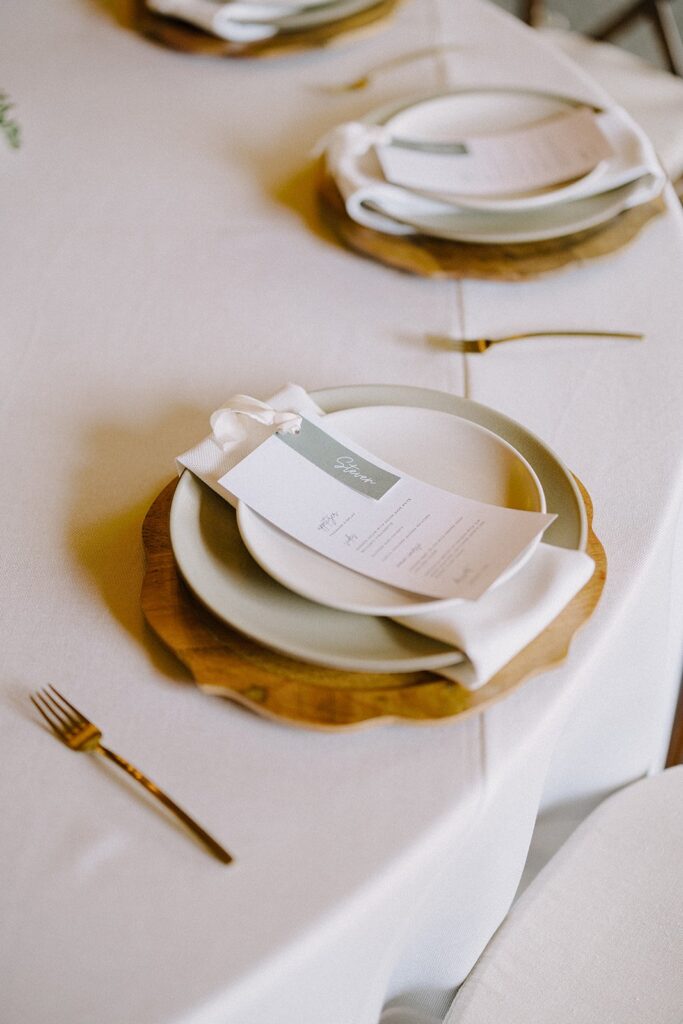 Elegant table setting with a layered white plate, napkin, and menu on a wooden charging plate, arranged on a white tablecloth with gold cutlery.