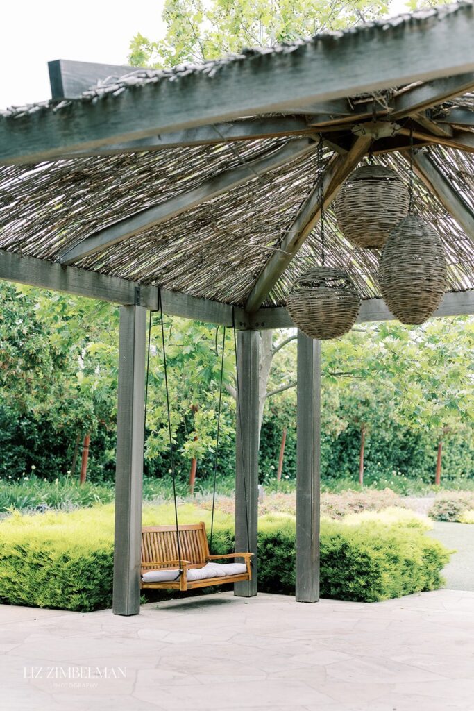 A wooden swing under a pergola with woven roof and hanging lanterns, surrounded by greenery and a stone patio.