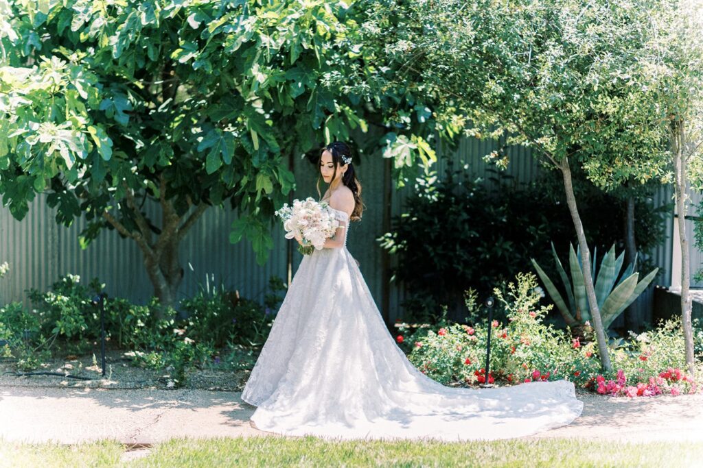 Bride in white gown holding a bouquet, standing on a garden path beneath lush trees, with flowers and greenery in the background.