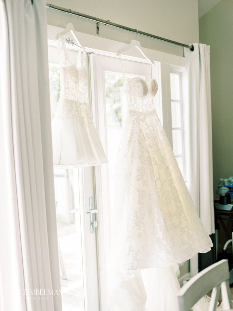 Two white lace bridal dresses hang in front of a sunny window with white curtains.