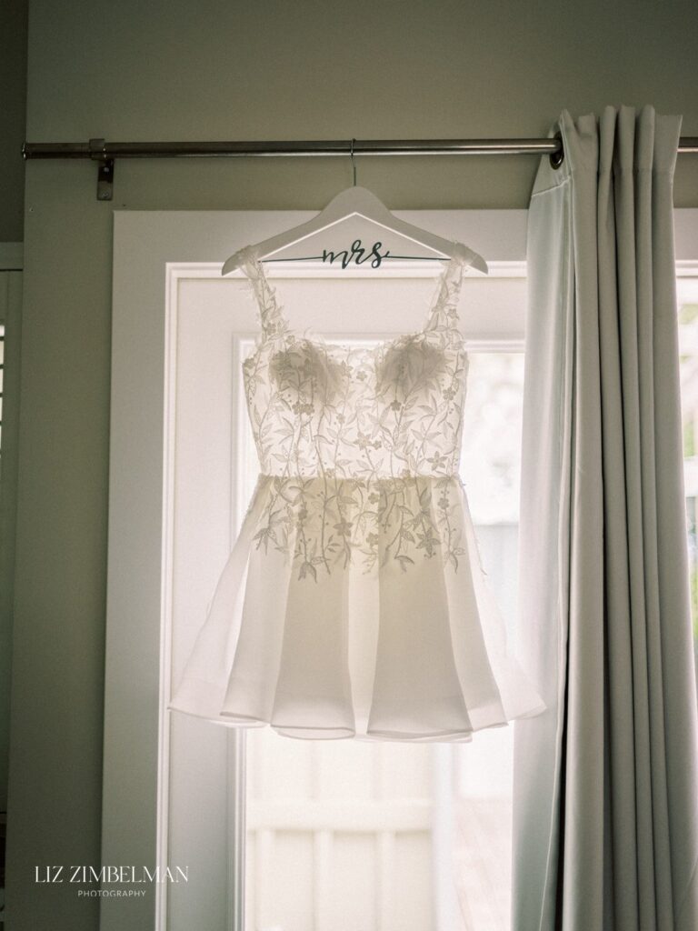 A white bridal dress with floral embroidery is hanging on a hanger labeled "mrs" near a window.