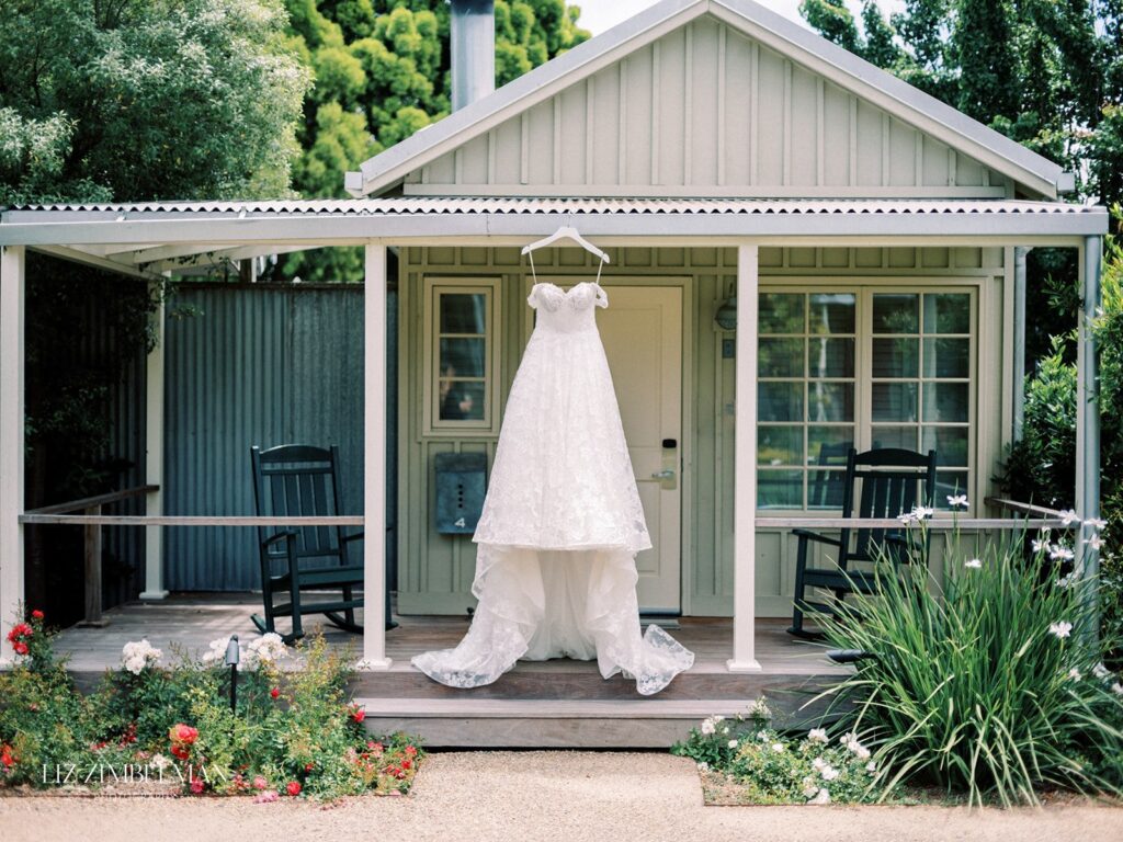 A wedding dress hangs on the porch of a small house with a white facade. Two rocking chairs are positioned on either side, and there are flowers and greenery in the foreground.