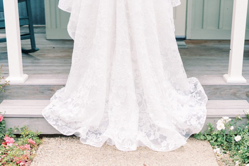 A white lace wedding dress train drapes over wooden steps, surrounded by small pink and white flowers.
