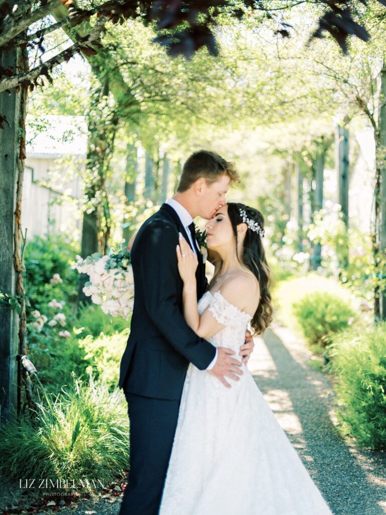 A bride and groom share a kiss on a path surrounded by greenery, with sunlight filtering through the trees.