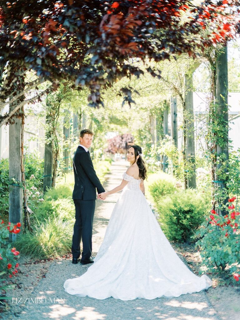 A bride and groom hold hands under a leafy pergola. The bride wears a white gown, and the groom is in a black suit. The path is lined with greenery and red flowers.