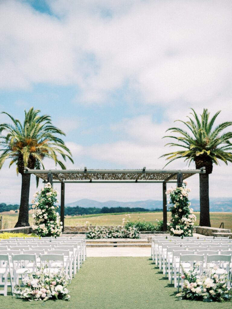 Outdoor wedding setup with white chairs facing a pergola adorned with flowers. Two palm trees frame the scene under a partly cloudy sky.