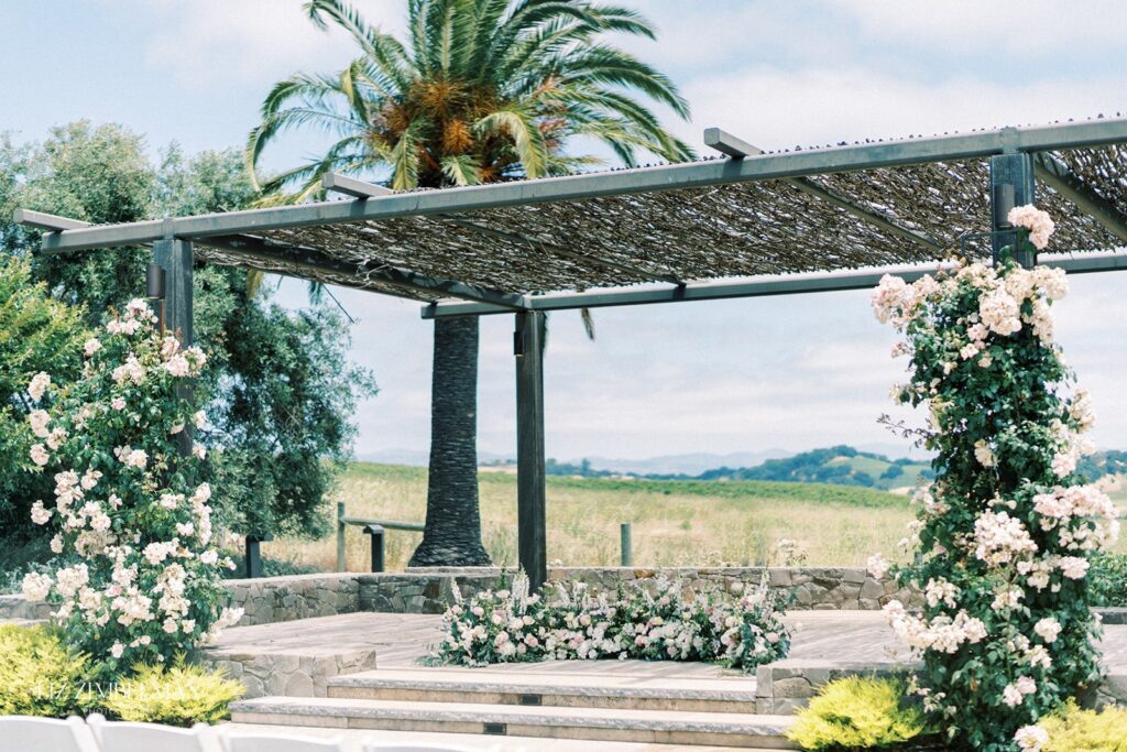 Outdoor wedding altar with a pergola covered in woven branches, adorned with pink and white flowers. Palm and floral surroundings with a scenic countryside backdrop.