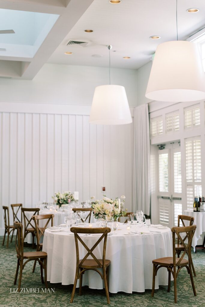 Elegant dining room with round tables set for an event, featuring white tablecloths, floral centerpieces, wooden chairs, and large overhead pendant lights.