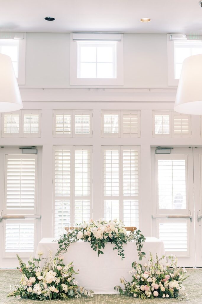 Table decorated with white and pink flowers, set in a bright room with large shuttered windows.