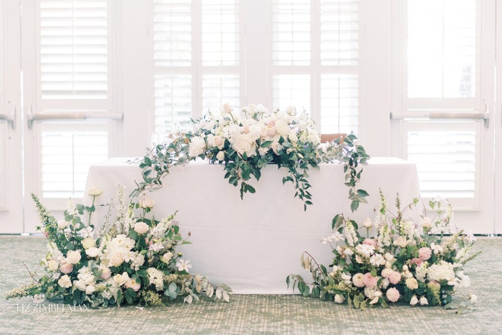 Elegant floral arrangement in white and pastel colors decorates a table covered with a white cloth, set against large windows with shutters.