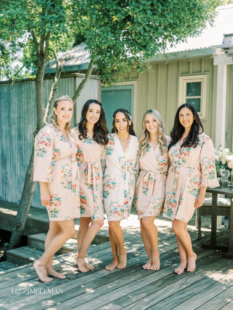 Five women in floral robes stand together on a wooden deck near a tree and a small building, smiling at the camera.