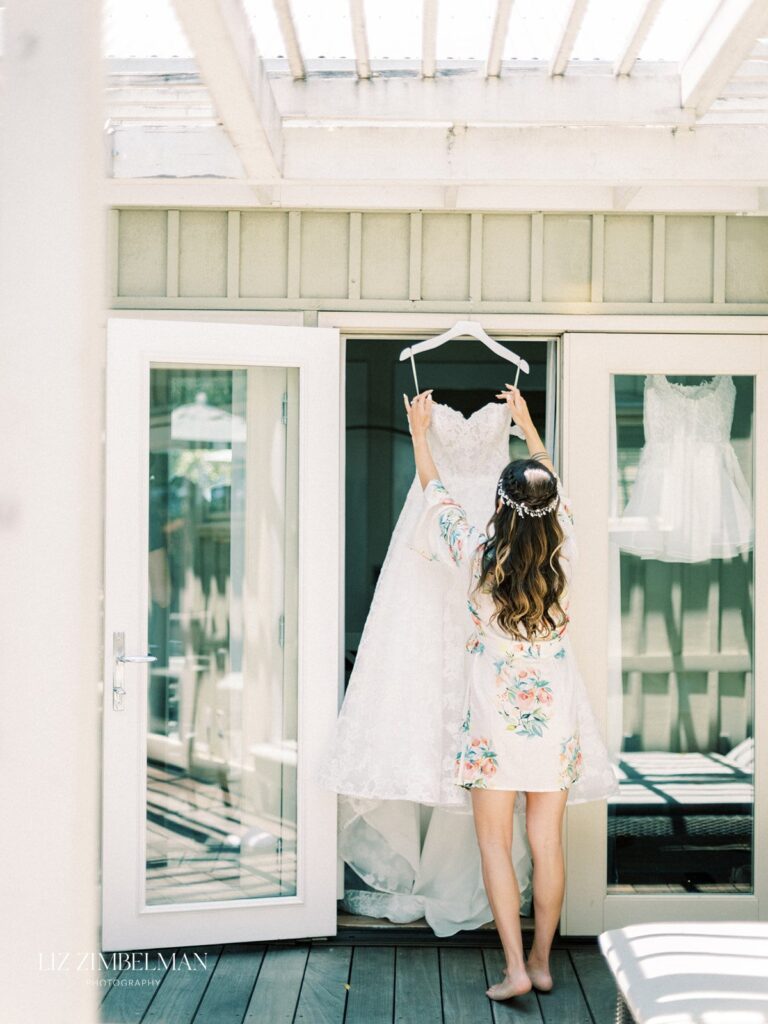 A woman in a floral robe holds a white wedding dress on a hanger in front of double doors on a wooden deck.