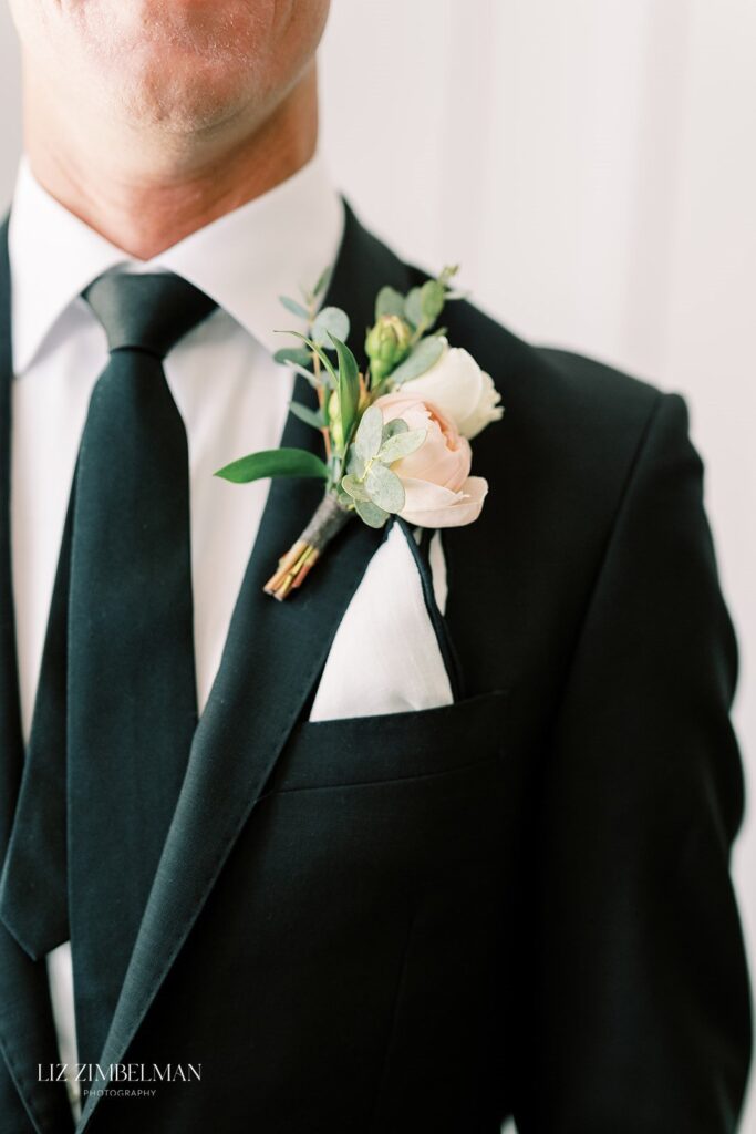 A person in a black suit and black tie, wearing a white shirt. A boutonniere with white and pink flowers is pinned to the lapel.