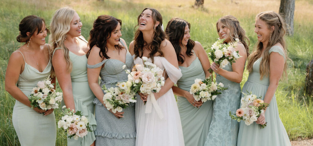A bride in a white dress stands with six bridesmaids in light teal dresses, all holding bouquets, outdoors in a wooded area.