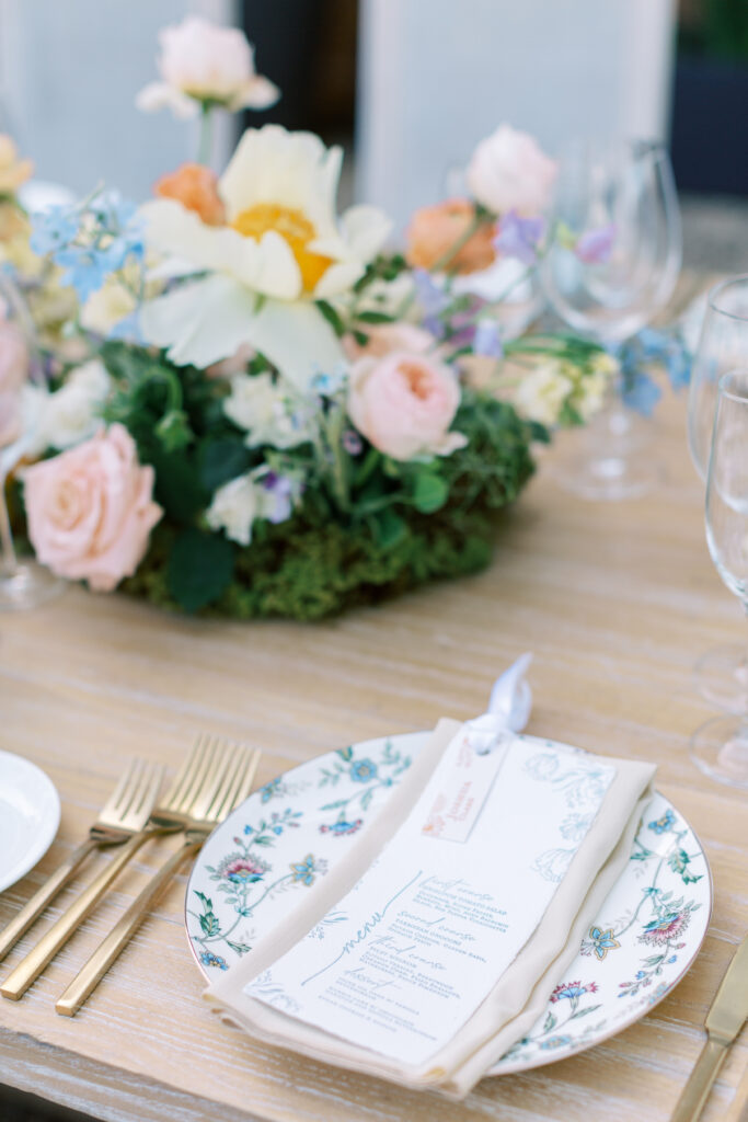 A floral plate setting with a printed menu and beige napkin, gold cutlery, and a centerpiece of pastel flowers on a wooden table.