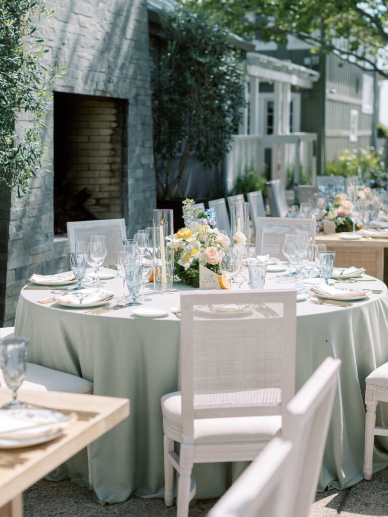 Outdoor dining area with round tables covered in light green tablecloths, set with glassware, plates, and floral centerpieces. White chairs surround the tables, near a stone fireplace.