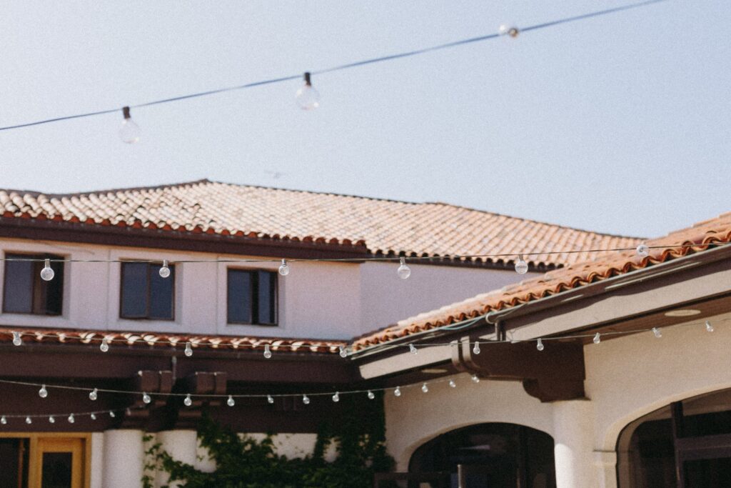 A courtyard with string lights hanging above, surrounded by buildings with terracotta roofs and white walls under a clear blue sky.