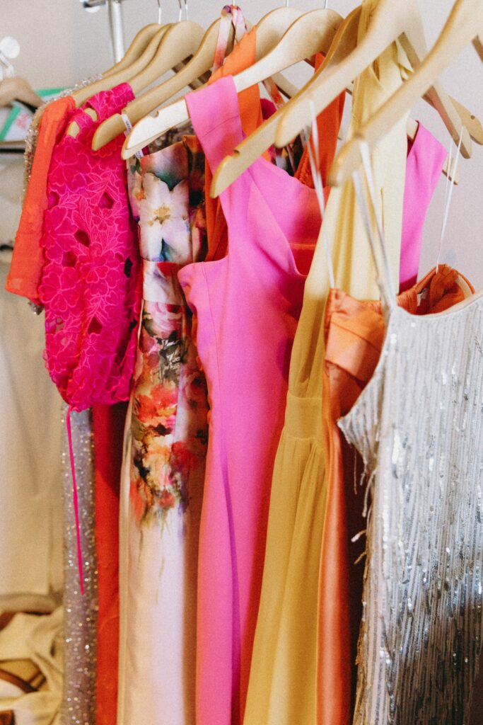 A selection of colorful dresses, including pink, floral, orange, and a silver sequin dress, hanging on wooden hangers in a closet.