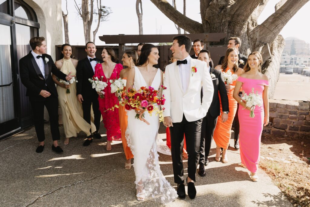 A wedding party walks outdoors, featuring a bride in a white dress and a groom in a white jacket. Bridesmaids and groomsmen follow, wearing colorful dresses and black suits.
