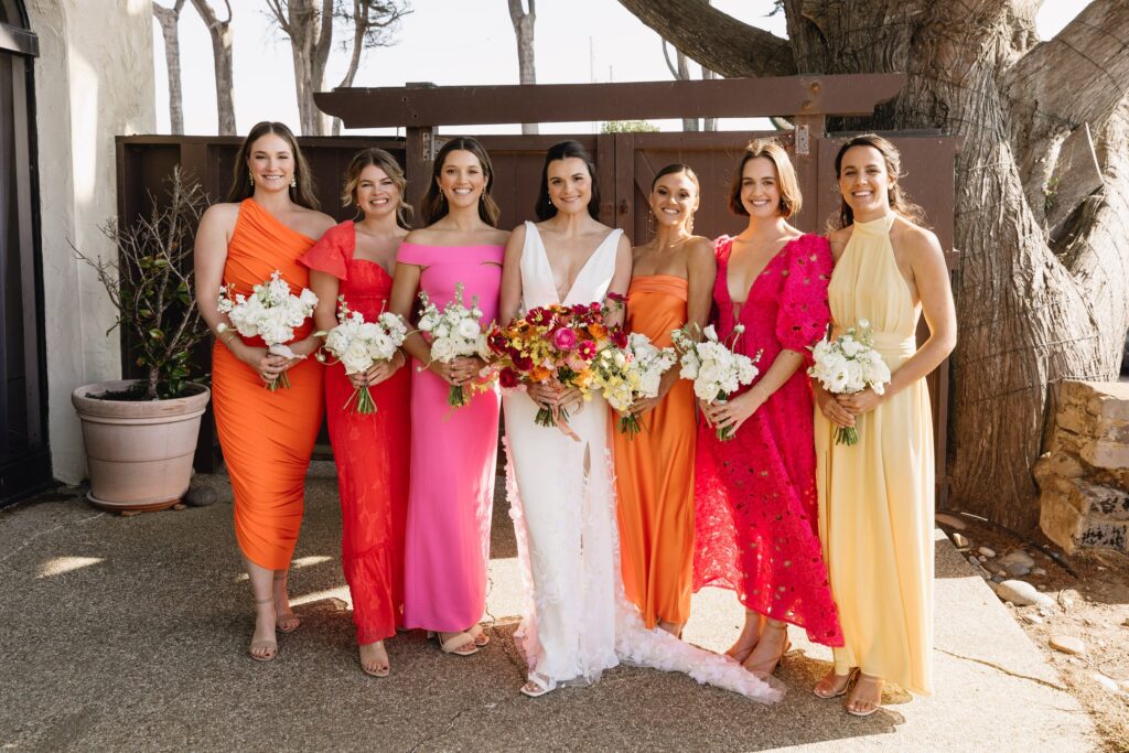 Seven women in colorful dresses stand side by side outdoors, holding bouquets.
