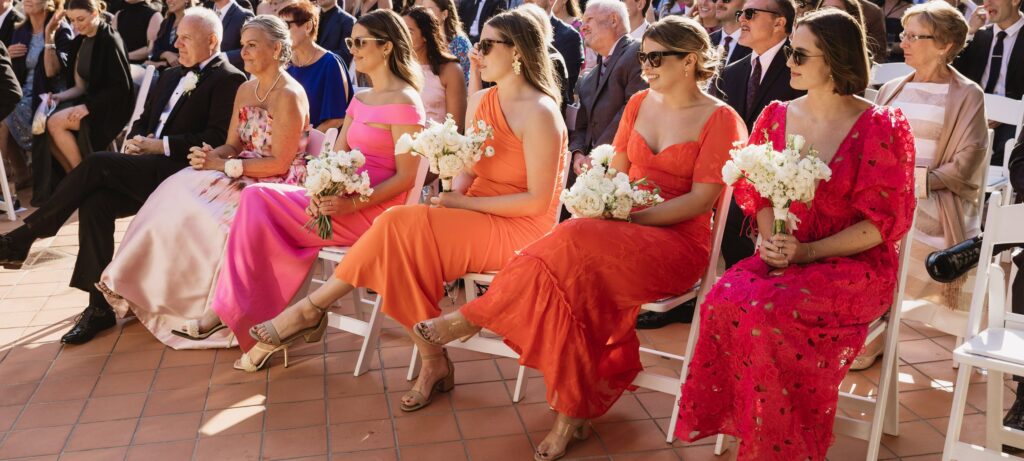 A group of people seated outdoors at a formal event, with women in bright dresses holding white floral bouquets, and string lights overhead.