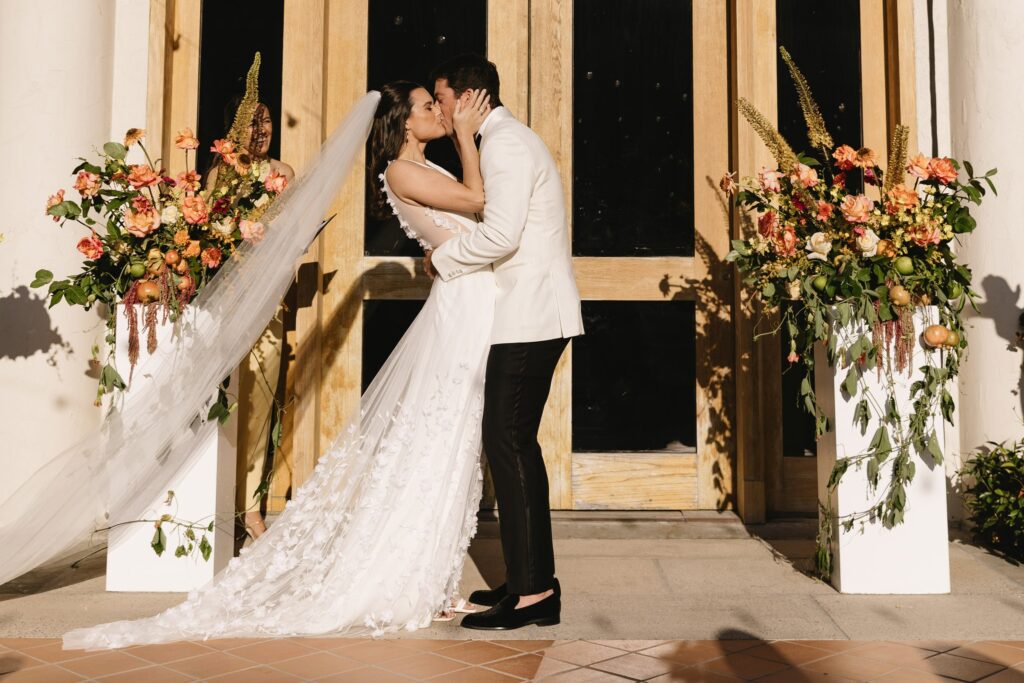 A bride and groom kiss in front of large wooden doors, flanked by floral arrangements with orange and pink flowers.