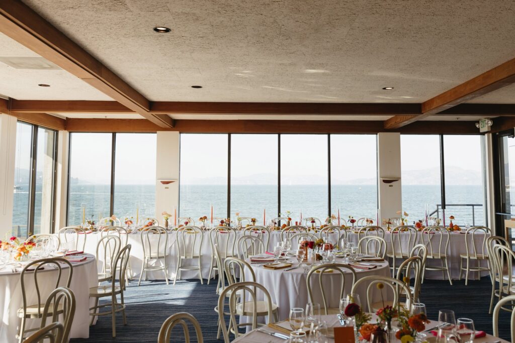 A dining room with round tables set for an event, each with white tablecloths and floral centerpieces. Large windows offer a view of the sea.