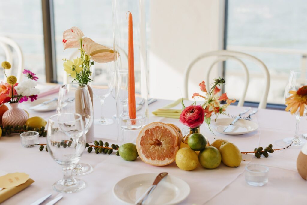 Elegant table setting with fruit centerpiece, flowers, glasses, and an orange candle on a white tablecloth. Scenic view through large windows.