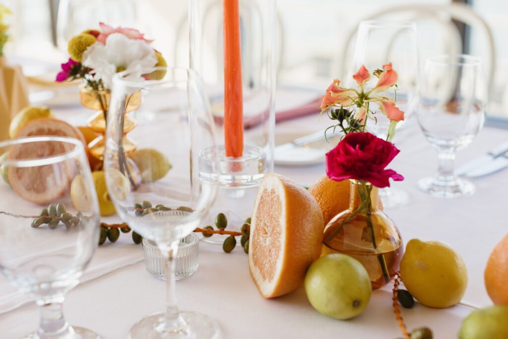 Elegant table setting with empty wine glasses, sliced grapefruit, whole pears, flowers in vases, and a tall orange candle on a white tablecloth.