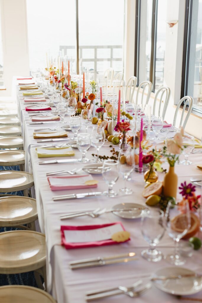 A long, formal dining table set with white tablecloths, arranged with glasses, cutlery, colorful flowers, and candles, surrounded by white chairs.
