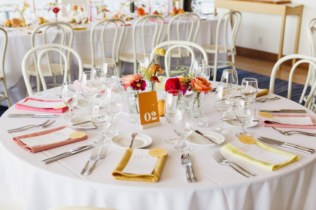 A round table set for a formal event with glasses, cutlery, and colorful napkins. Centerpiece includes red and orange flowers. A table number card marked "02" is displayed.